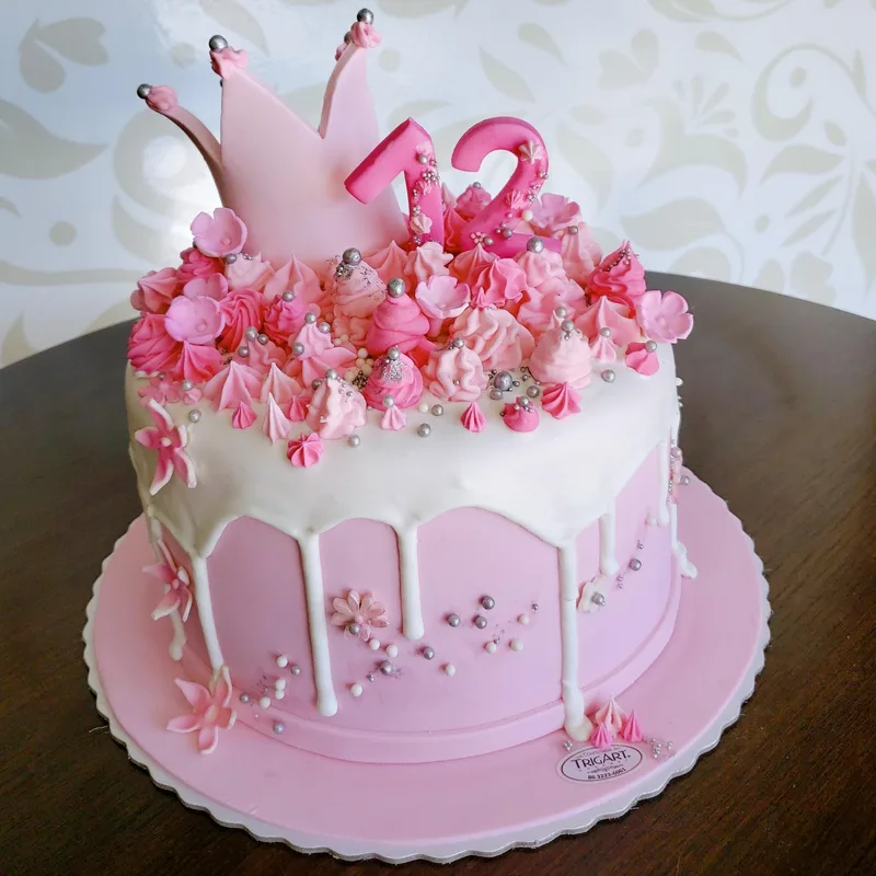 Bolo rosa com cobertura branca escorrendo, merengues, flores de pasta e coroa rosa com o número 12 no topo.