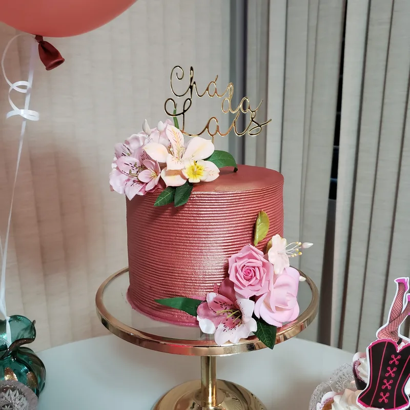Bolo rosé com textura em linhas, flores de açúcar rosadas e letreiro dourado no topo, sobre suporte dourado.