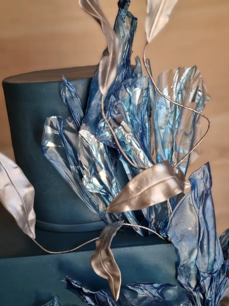 Detalhe de bolo azul-petróleo com folhas de pasta metalizadas em azul e prata.