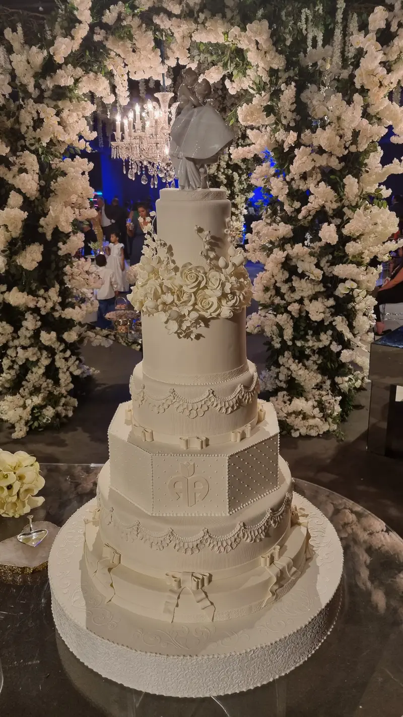 Bolo de casamento de seis andares, bege, com relevos e monograma, diante de um arco de flores brancas.