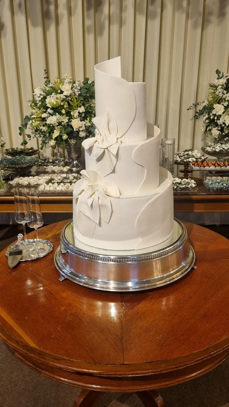Bolo de casamento de quatro andares, branco, com laço e ondas de pasta em linhas limpas, sobre suporte prateado.