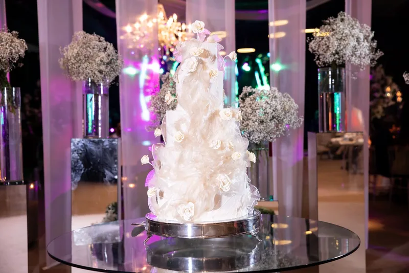 Bolo de casamento alto, coberto por drapeado translúcido e rosas brancas, em cenário iluminado com gipsófila.
