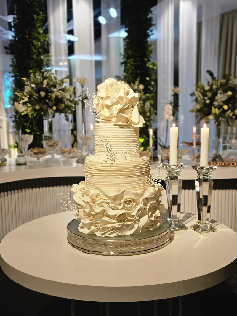 Bolo de casamento de quatro andares em creme, com camadas franzidas e uma grande flor de pasta no topo.