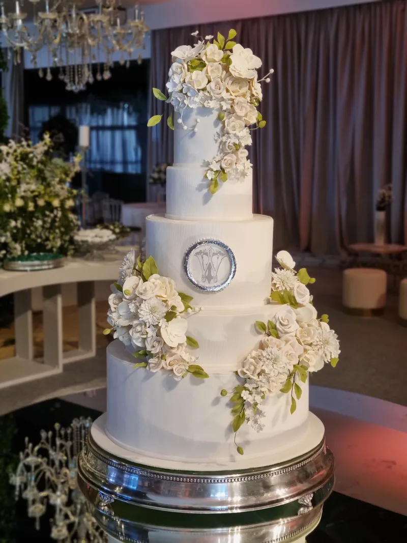 Bolo de casamento de quatro andares, branco, com cascata de flores de açúcar brancas e folhas verdes e medalhão prateado.