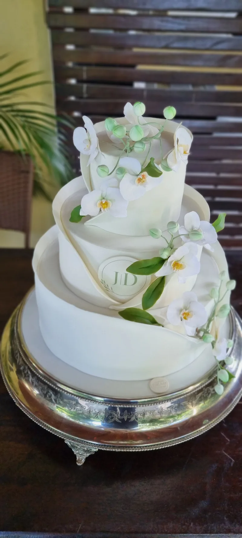 Bolo de casamento de três andares com camadas inclinadas e orquídeas brancas, sobre suporte prateado.