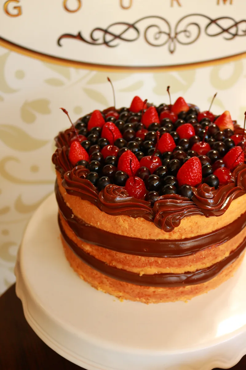 Bolo naked de massa dourada e camadas de chocolate, com frutas vermelhas e cerejas no topo.