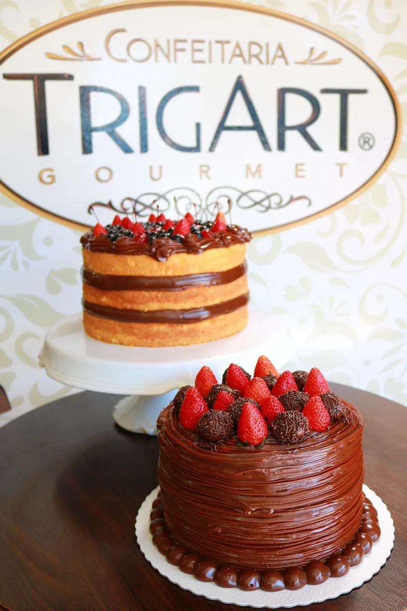 Dois bolos, um naked de chocolate e um coberto de ganache com morangos, diante do letreiro da Trigart Gourmet.