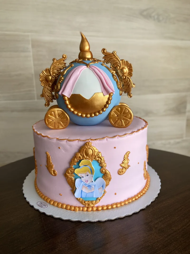 Bolo infantil de dois andares, rosa, com carruagem azul e dourada de pasta americana no topo e medalhão de princesa na lateral.