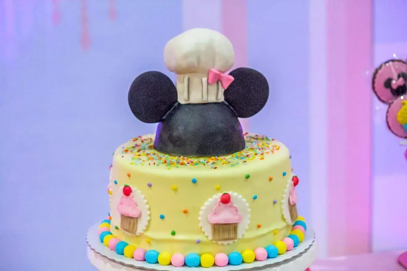 Bolo infantil amarelo com confeitos coloridos e, no topo, orelhas de Minnie com chapéu de cozinheira.