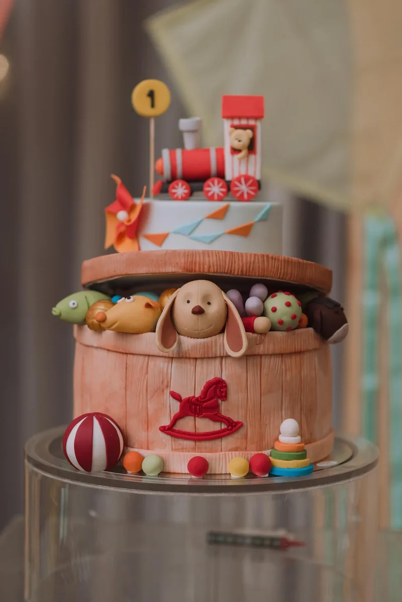 Bolo infantil em forma de baú de brinquedos com cachorrinho, cavalinho de balanço e trem vermelho no topo.