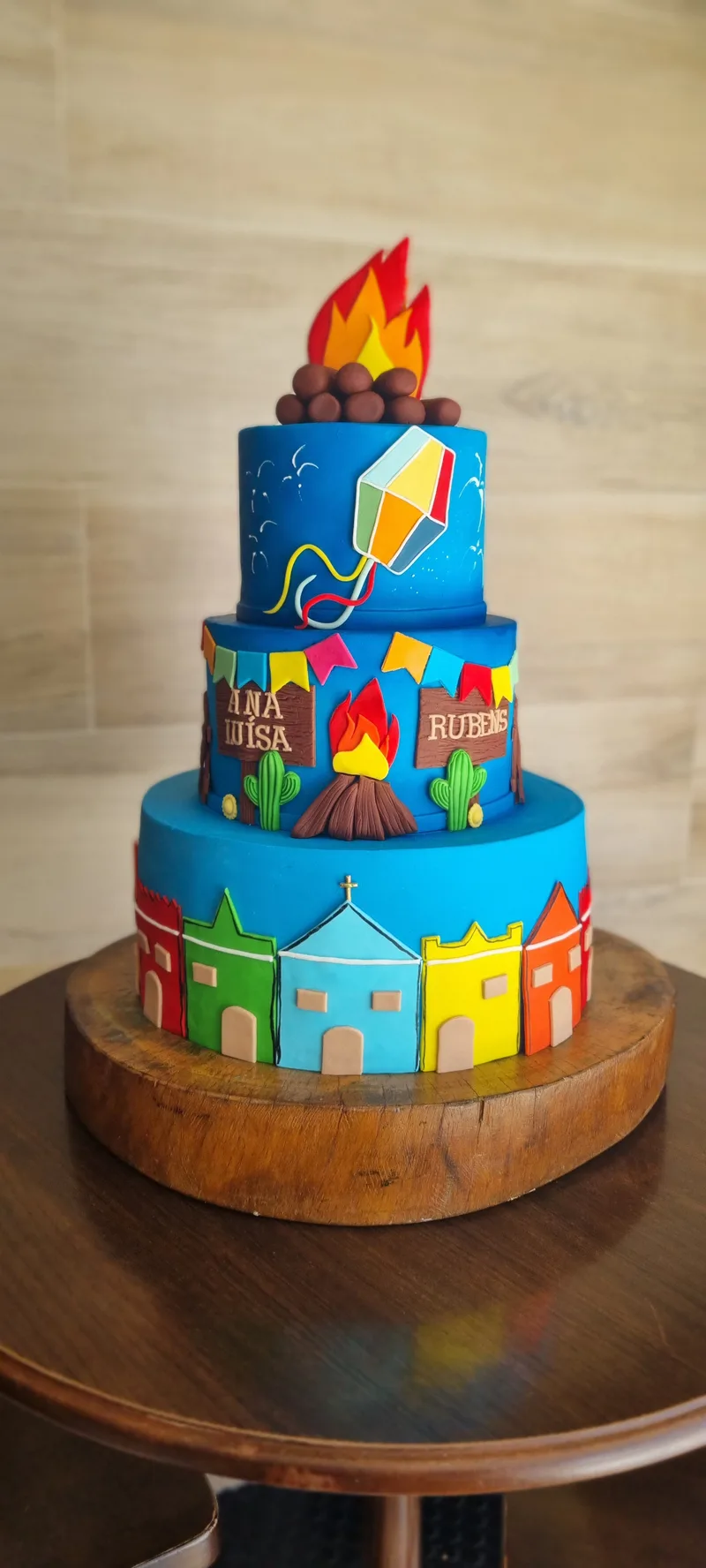 Bolo de festa junina de três andares em azul, com casinhas coloridas, bandeirinhas, fogueira e balão no topo.