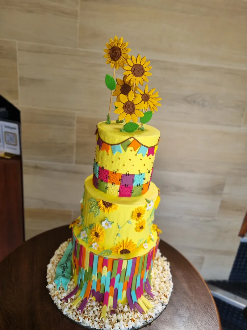 Bolo de festa junina de três andares em amarelo, com girassóis, patchwork colorido e franjas na base.