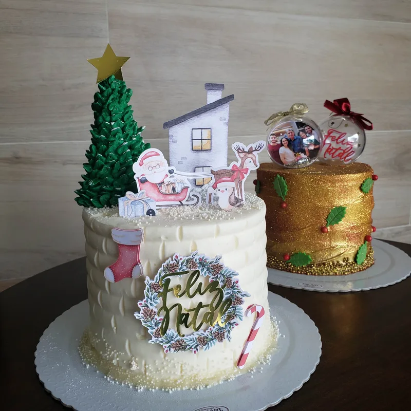 Dois bolos de Natal lado a lado, um branco com árvore, casa e Papai Noel e outro dourado com esferas e azevinho.