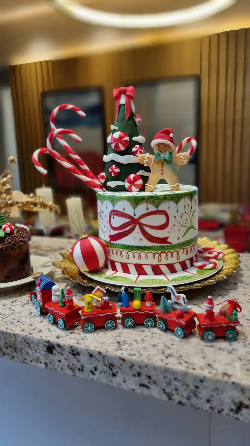 Bolo de Natal listrado em vermelho, branco e verde, com laço, árvore, boneco de biscoito e bengalas, e trenzinho de brinquedo na base.